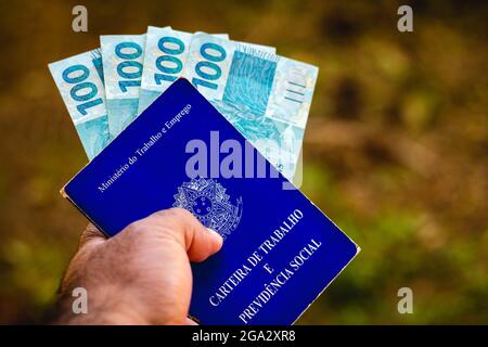 In this photo illustration, a man displays his Work and Social Security Card of Brazil with one hundred reals bills.It is the document that guarantees access to the rights of the Brazilian worker. Stock Photo