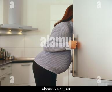 A pregnant woman stands with the door open to the refrigerator and looking in; Germany Stock Photo