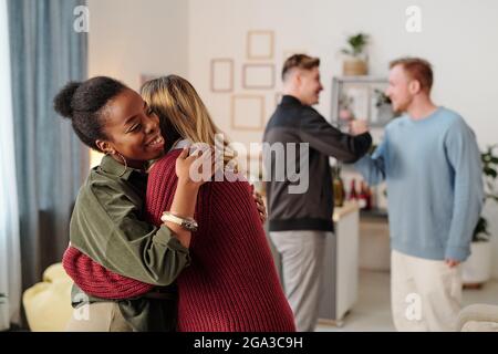 Happy young African woman in smart casualwear giving hug to guy in blue pullover in front of camera against their friends embracing and talking Stock Photo