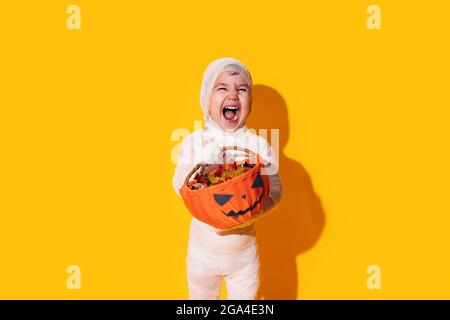 Child in mummy costume holding basket of chocolates in front of yellow background. Stock Photo