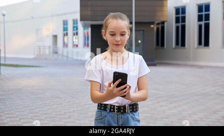 Schoolgirl walks along street. Addicted to smartphone teenage girl looks at display and types on move walking against large city building closeup Stock Photo