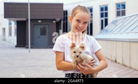 Young lady poses with dog on street. Teenage girl in white t-shirt holds small puppy in arms and looks straight smiling by city buildings close view Stock Photo