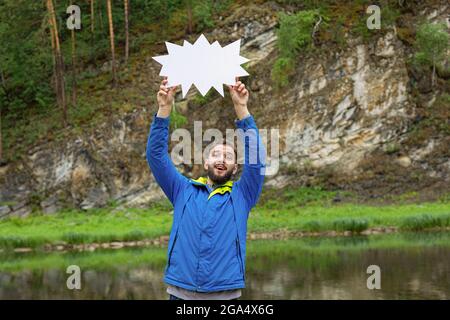 Smiling young man with beard in jacket holding blank speech bubble above your head against background of natural landscape. having idea, want to speak Stock Photo