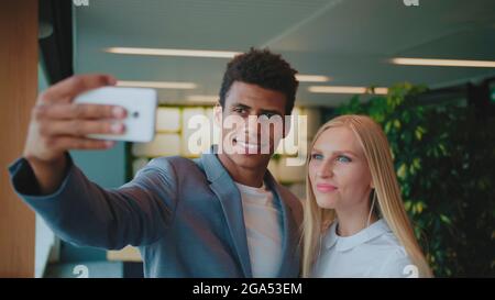 Cheerful black man with laughing blond woman taking selfie with smartphone in modern office having fun. Stock Photo