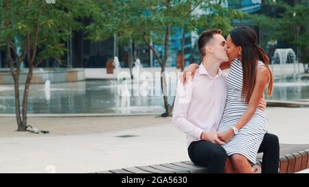 Two mixed-race lovers sitting on a bench and kissing. They having a date in a beautiful place in the city center. Stock Photo