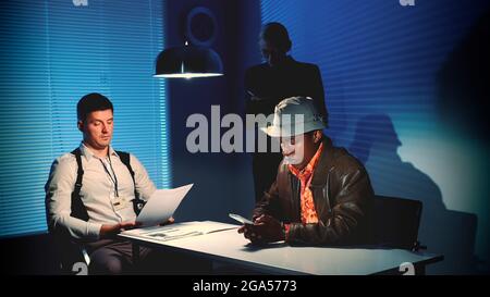 Multiracial desperate criminal writing last message on smartphone in interrogation room. Police detective pulling the phone from the accused man. Stock Photo