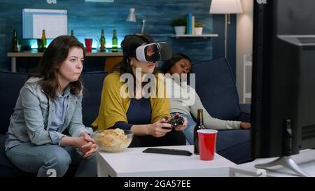 African woman experiencing virtual reality playing video games with vr headset and joystick in home living room while friends are cheering up for her. Mixed race group of people hanging out having fun Stock Photo