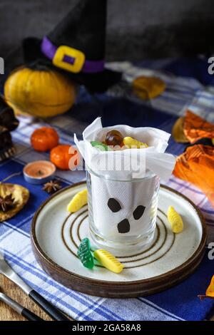 Halloween party. White ghost crafts scary face in glass on a plate and a festive Halloween table setting with pumpkins. Stock Photo
