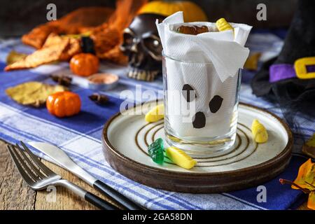 Halloween party. White ghost crafts scary face in glass on a plate and a festive Halloween table setting with pumpkins. Stock Photo