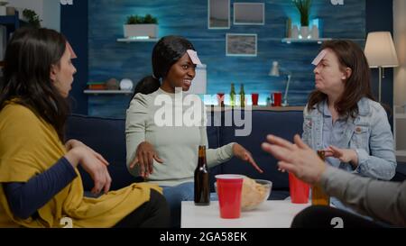 Afro american woman having attaching sticky notes on forehead playing guess who game with friends during creative party. Mixed race people having fun together while sitting on sofa late at night Stock Photo
