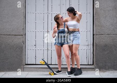 Two cool women hugging and looking to each other with a skateboard. Two female friends posing at street. Stock Photo