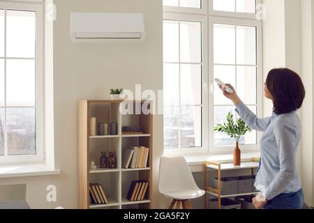 Woman standing in living room switches modern air conditioner with remote control Stock Photo