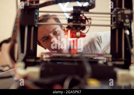 Focused young man watching modern 3D printer creating red plastic model in studio Stock Photo
