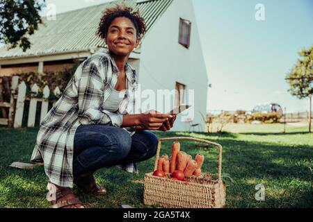 Mixed race female teen crouching down in garden next to basket of fresh vegetables researching information on digital tablet  Stock Photo
