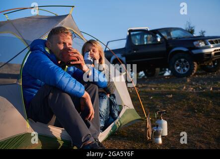 Couple of tourists drinking freshly brewed tea sitting in tent set up on lawn at sunny weather in early autumn. On the background black SUV parked on Stock Photo