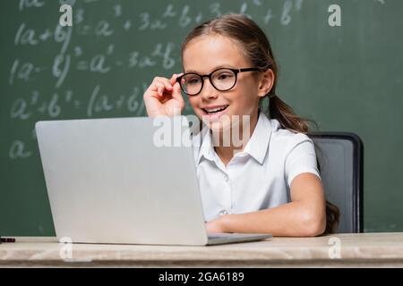 positive schoolkid adjusting eyeglasses near laptop and chalkboard on blurred background Stock Photo