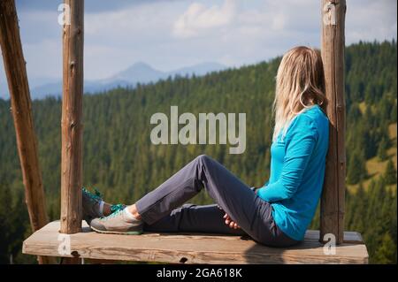 Side view of young blonde girl resting alone in mountains, sitting on seat of large wooden swing and enjoying breathtaking view of the mountain beskids. Stock Photo