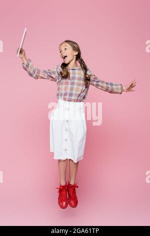 astonished child in trendy clothes levitating with digital tablet on pink background Stock Photo
