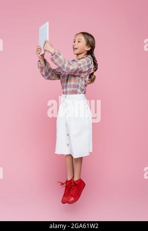 excited girl in stylish clothes levitating with digital tablet on pink background Stock Photo