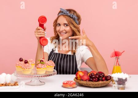 Portrait of blonde woman in white T-shirt and apron sitting at table surrounded with pastry and fruits and holding handset, showing call me gesture. I Stock Photo