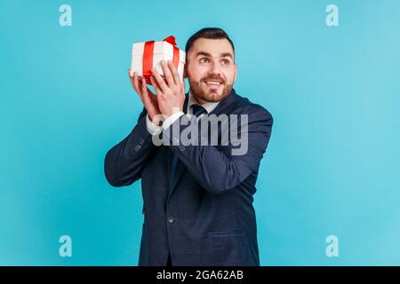 Curious dreamy bearded man in elegant suit shaking gift box trying to guess what is inside, wondered with present, surprises and bonuses. Indoor studi Stock Photo