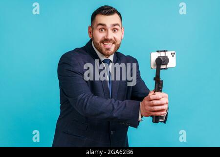 Portrait of happy positive bearded businessman wearing official style suit streaming online, using smart phone and steadicam, broadcasting live stream Stock Photo