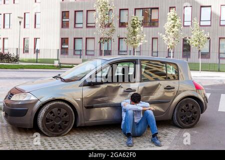 Man in despair wearing casual clothing sitting on ground with tilted head near broken car, damaged automobile with scratches and dents on doors, being Stock Photo