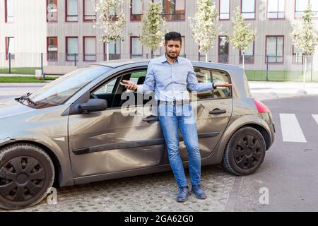 Young adult bearded man with sad expression standing with raised arms and looking at camera, being confused after car accident, broken auto with dents Stock Photo