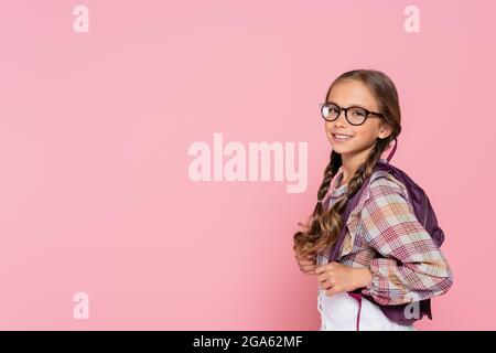 Preteen schoolgirl in eyeglasses and backpack smiling at camera isolated on pink Stock Photo