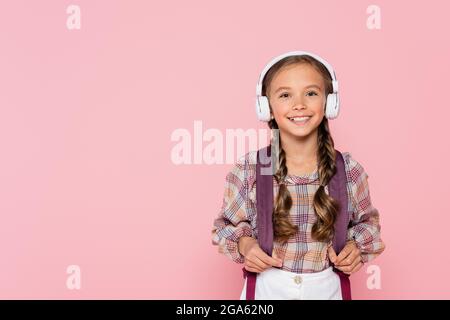 Smiling schoolgirl in headphones holding backpack isolated on pink Stock Photo