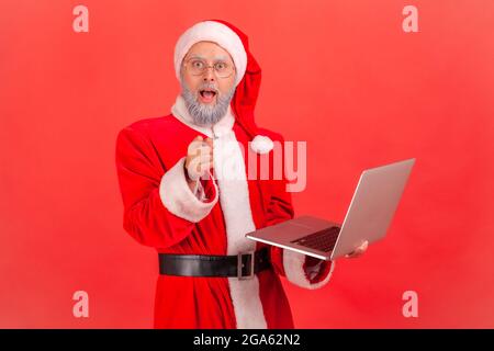 Shocked elderly man with gray beard wearing santa claus costume holding laptop in hands and pointing to camera with big eyes and astonishment. Indoor Stock Photo