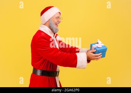 Side view of elderly man with gray beard wearing santa claus costume holding giving present box, greeting with Christmas and new year eve. Indoor stud Stock Photo