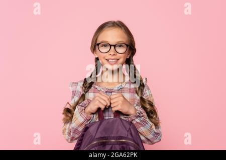 Schoolgirl in checkered shirt holding backpack and smiling at camera isolated on pink Stock Photo