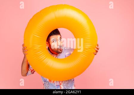 Portrait of funny bearded man on vacation, wearing casual clothing, holding orange rubber ring and looking away with amazement and interest. Indoor st Stock Photo