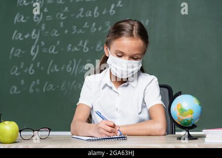 preteen schoolgirl in medical mask writing in notebook near globe, apple and blurred chalkboard Stock Photo