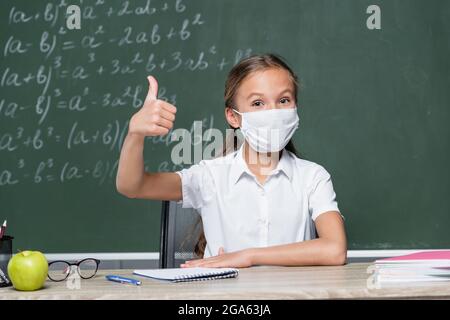 schoolkid in medical mask showing thumb up near apple, notebook and chalkboard on blurred background Stock Photo
