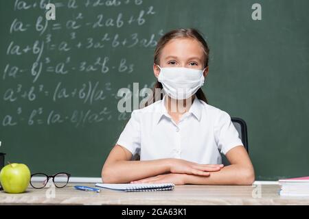 schoolkid in medical mask sitting at desk near apple, notebook, eyeglasses and blurred chalkboard Stock Photo
