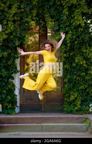 excited woman in yellow sports suit jumps and enjoy summer on green ivy background of overgrown greenhouse Stock Photo