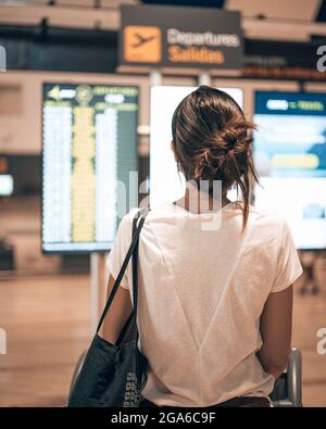 Caucasian woman going into the terminal airport during the covid pandemic loading her luggage into a trolley while looking at the departure screens Stock Photo