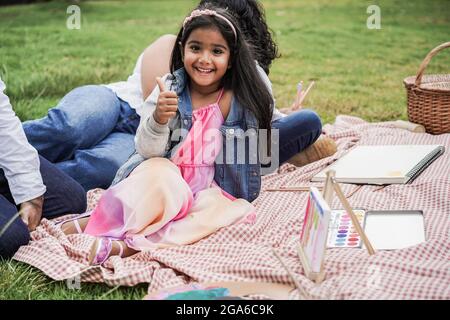 Happy indian family having fun painting with children outdoor at city park - Main focus on girl face Stock Photo
