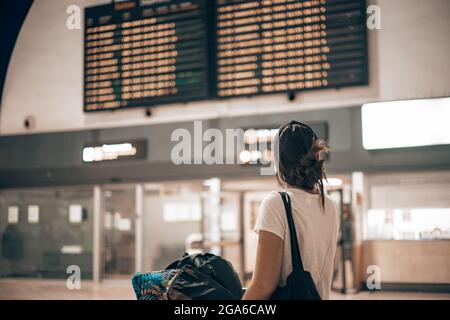 Caucasian woman walking through the airport terminal during the covid pandemic loading her luggage into a trolley looking at the big screens Stock Photo