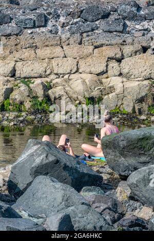 young couple sunbathing among the rocks on the beach at dunure harbour on the ayrshire coast in scotland. young couple both using smartphones. Stock Photo