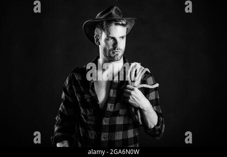 Lassoing on prairie. Man unshaven cowboy black background. Cowboy wearing hat hold rope. Lasso tool of American cowboy. Still used on ranches to Stock Photo