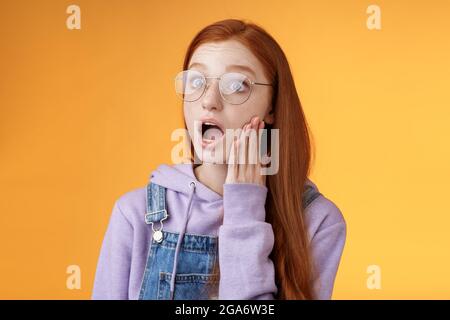 Shocked impressed speechless young redhead sensitive european girl glasses wearing hoodie dungarees drop jaw gasping astonished look left wide eyes Stock Photo