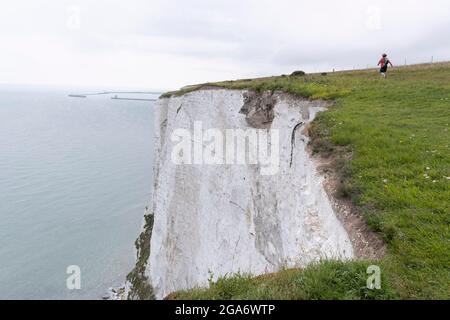 With Dover Dock's harbour wall in the distance, a lone woman walker strides on a clifftop footpath, near the edge of England's iconic White Cliffs, under threat from chalk and soil erosion, on 27th July, in St Margaret's at Cliffe, in Kent, England. Stock Photo