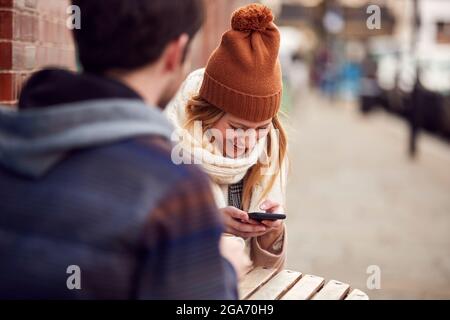 Couple On Date Sitting Outside Coffee Shop On High Street Using Mobile Phone Stock Photo