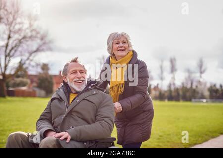 Senior Woman Pushing Senior Man In Wheelchair Outdoors In Fall Or Winter Park Stock Photo
