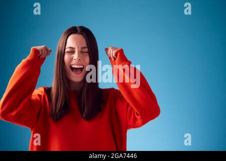 Excited Young Woman Celebrating And Pumping Fists In The Air Against Blue Studio Background Stock Photo