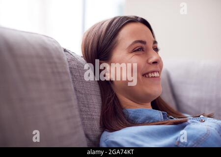 Smiling Young Woman At Home Lying On Sofa Watching TV And Laughing Stock Photo