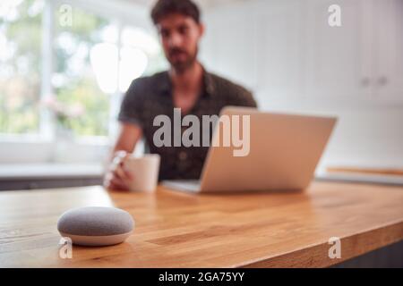 Man In Kitchen Using Digital Smart Speaker Whilst Working From Home With Laptop During Pandemic Stock Photo